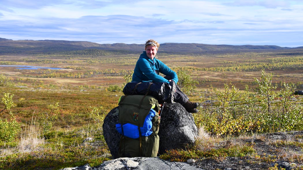 En vandrare sitter på en stor sten. Ett brett fjälllandskap öppnar sig i bakgrunden.